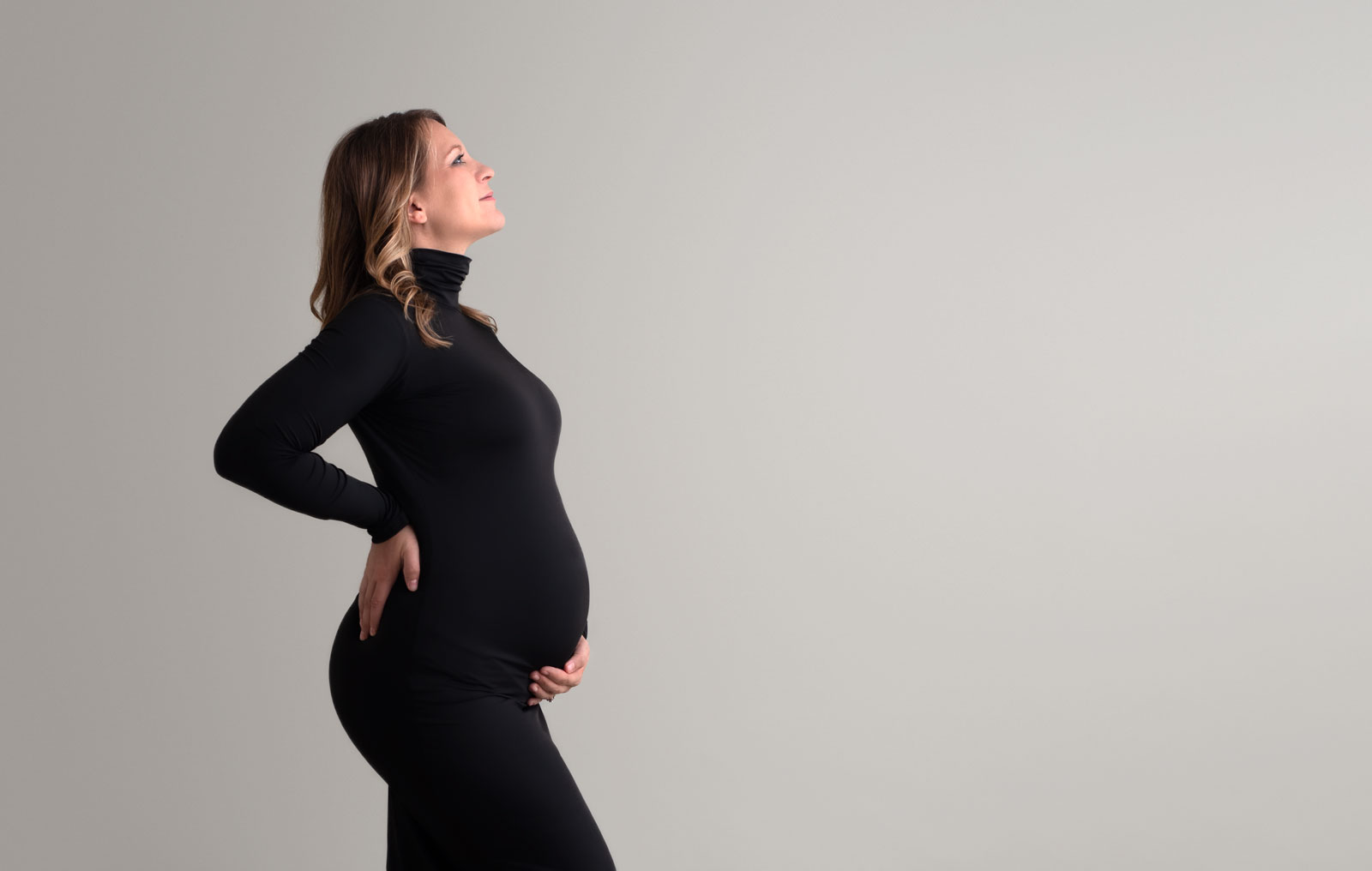 Mother to be in a black maternity dress stands proudly in a studio holding her bump dancing for birth