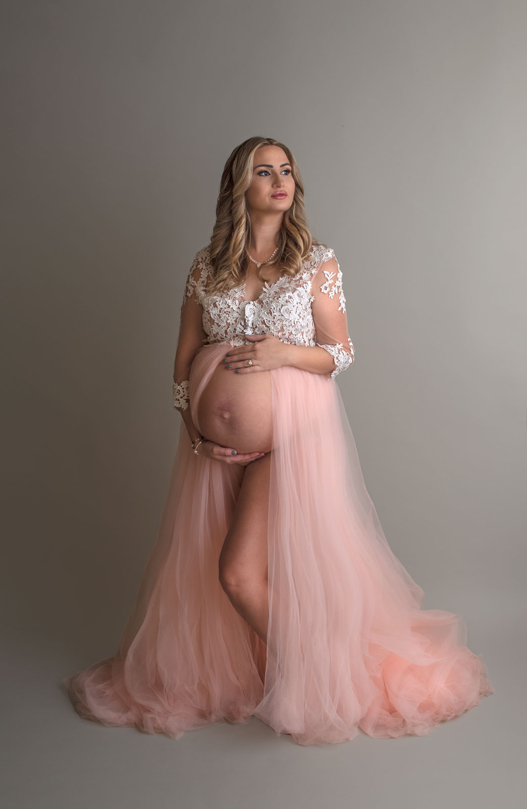 A mother to be stands in a studio wearing a maternity dress with white lace on top and pink velour draping around her bump St. louis maternity clothes
