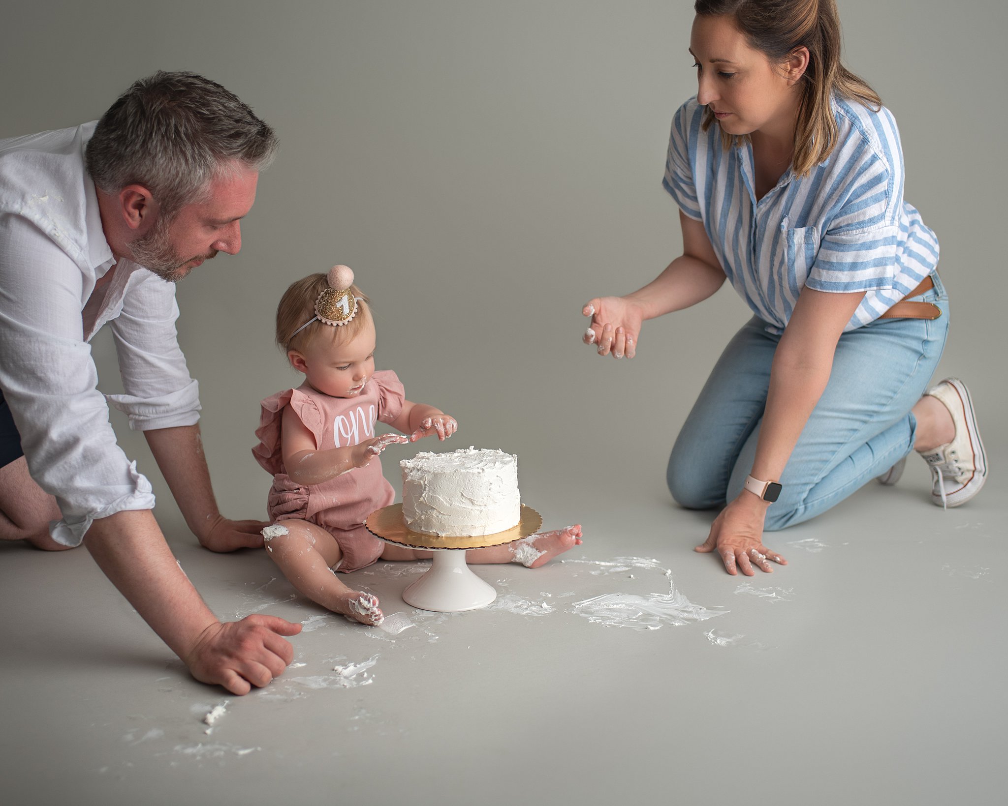 Mom and dad play with their one year old daughter on the floor of a studio as she plays in a white frosting birthday cake