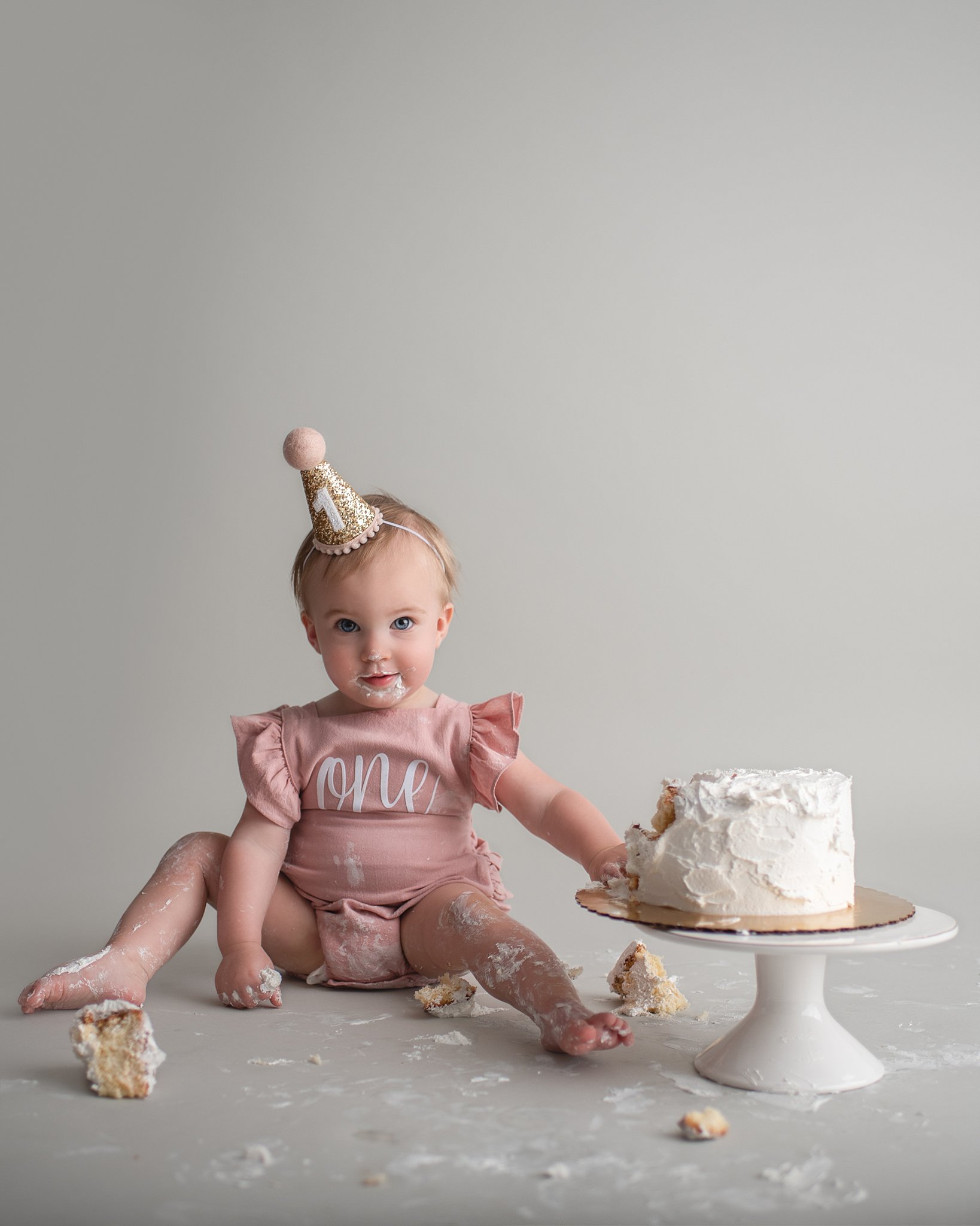 a toddler sits on the floor of a studio covered in cake that she smashed to celebrate her first birthday in a pink onesie just between friends st charles