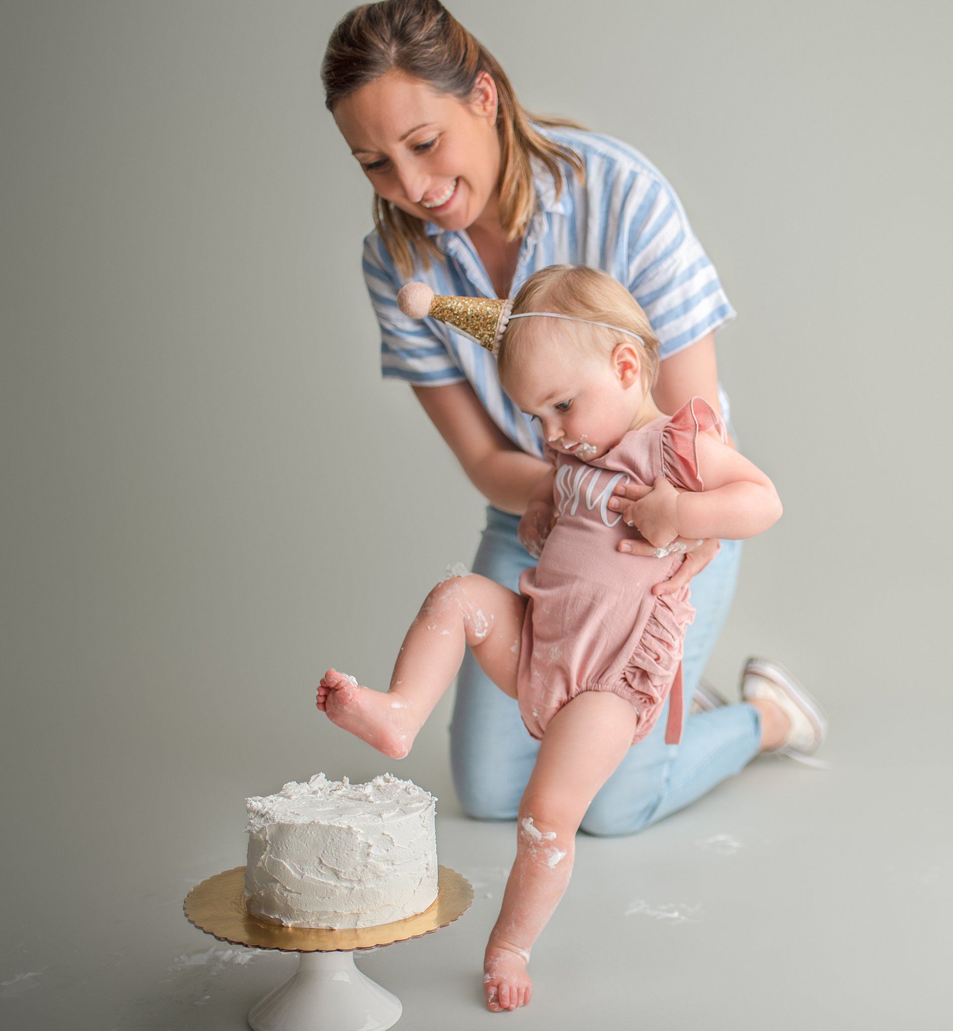 A mother holds her daughter up so she can kick her first birthday cake in a studio just between friends st charles