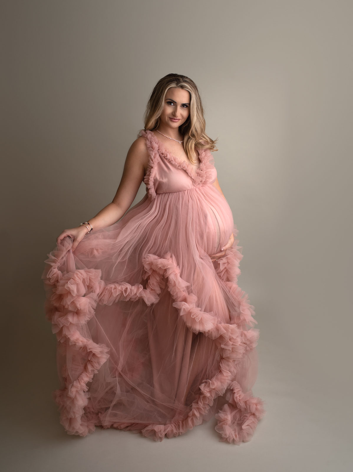 A mother to be stands in a pink maternity gown made of tule in a studio purple lotus doulas