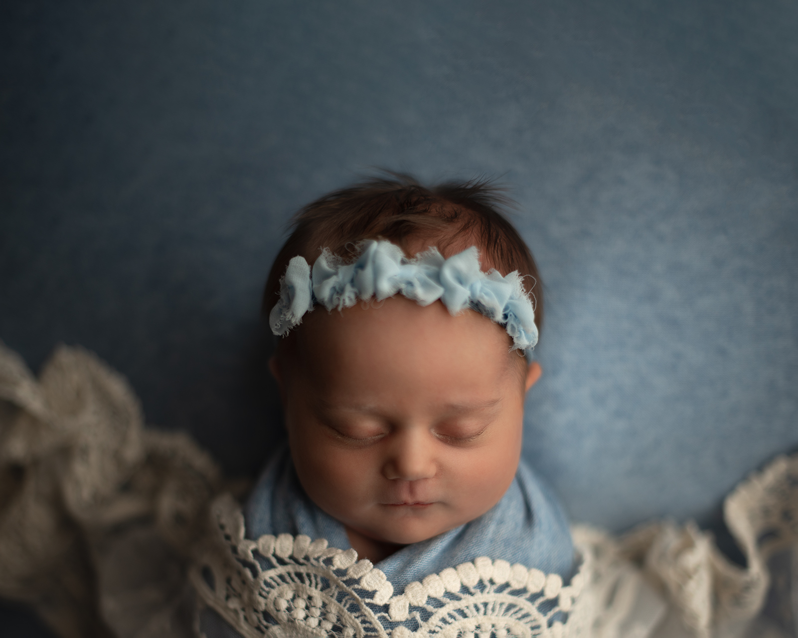 A newborn baby sleeps in a blue and lace swaddle wearing a blue headband st louis babysitters