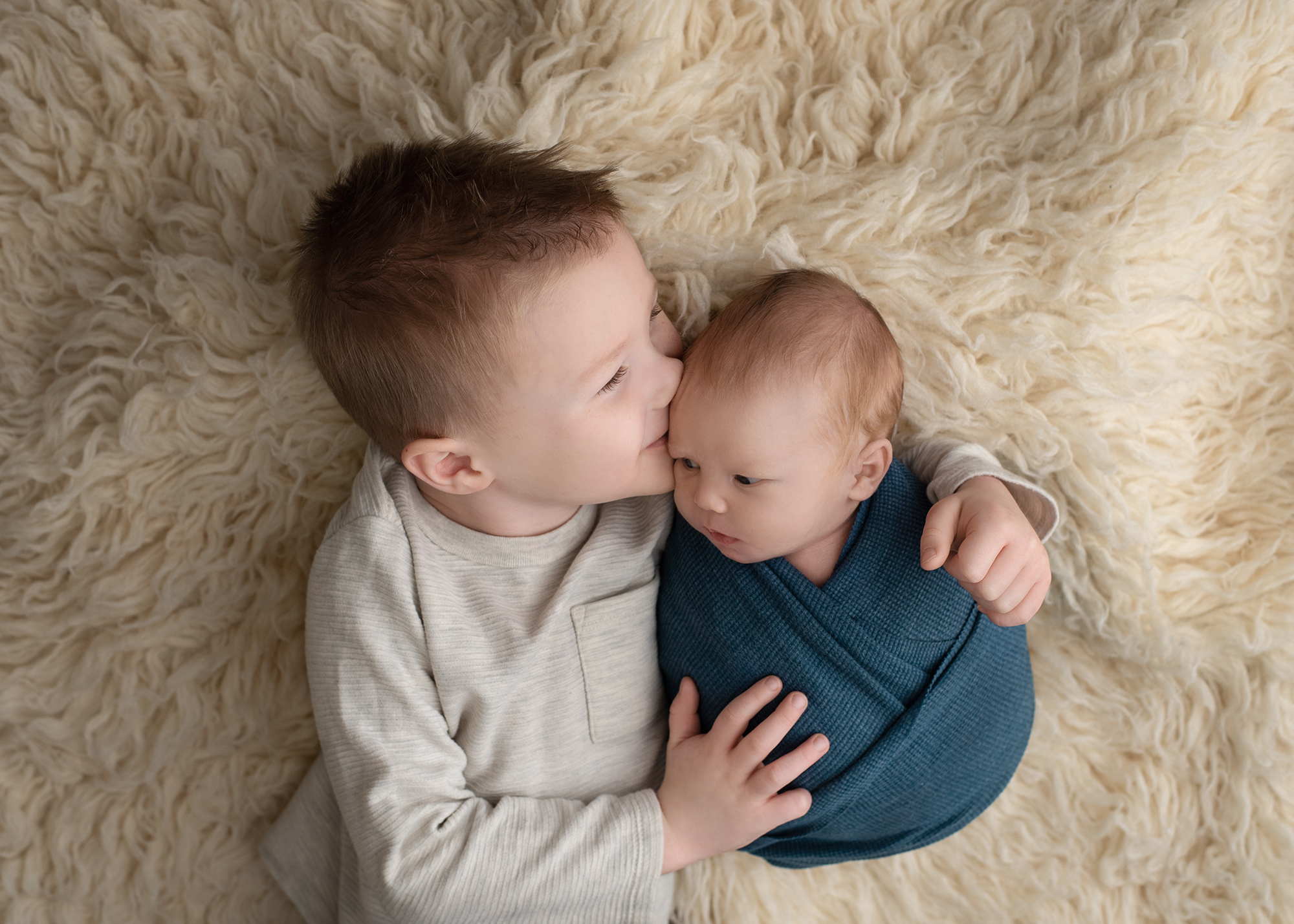A toddler boy kisses the head of his newborn baby brothe while laying on a furry blanket honeycomb st louis