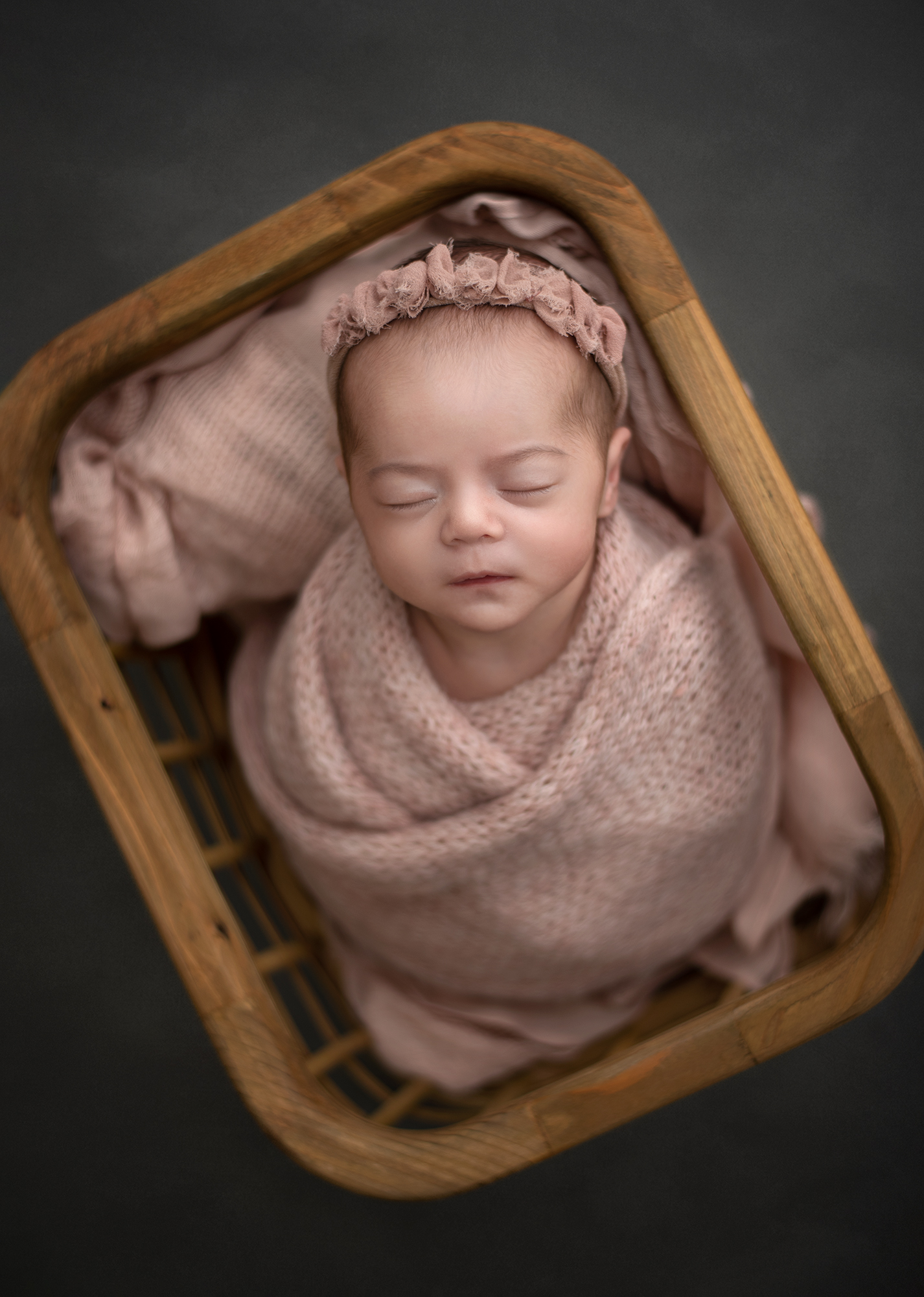 A newborn baby sleeps while swaddled in a pink knit blanket in a wooden basket