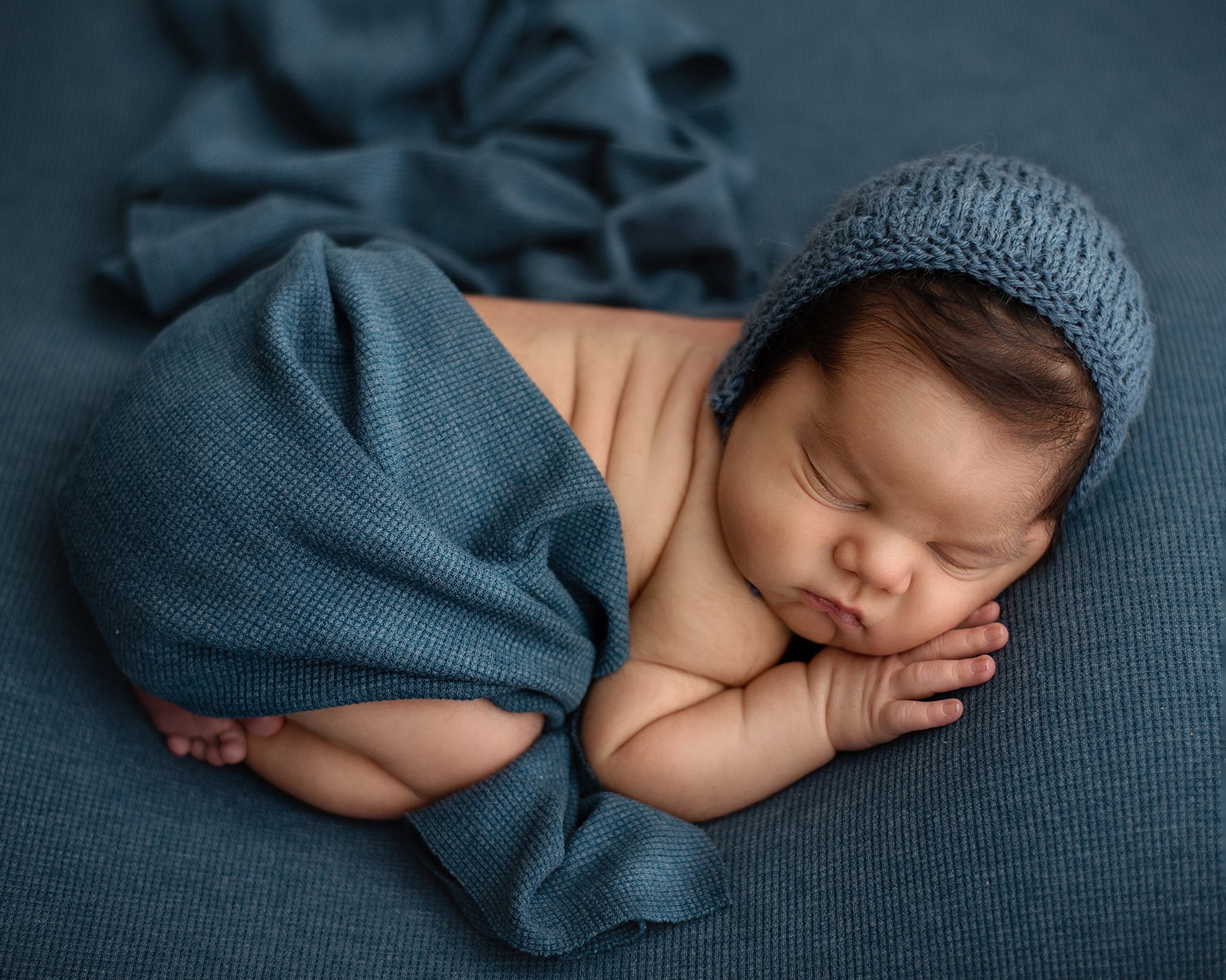 A newborn baby in a knit bonnet sleeps on a blue bed