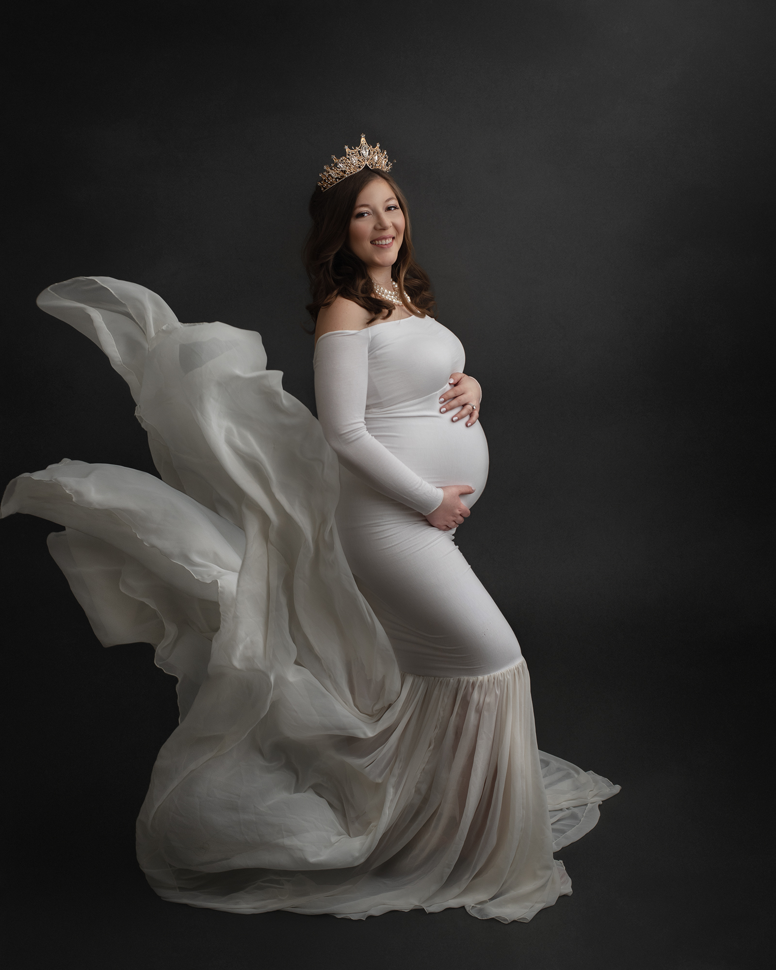 A mom to be in a crown and white maternity gown stands in a studio as the train flows in the wind behind her st louis baby shower venues