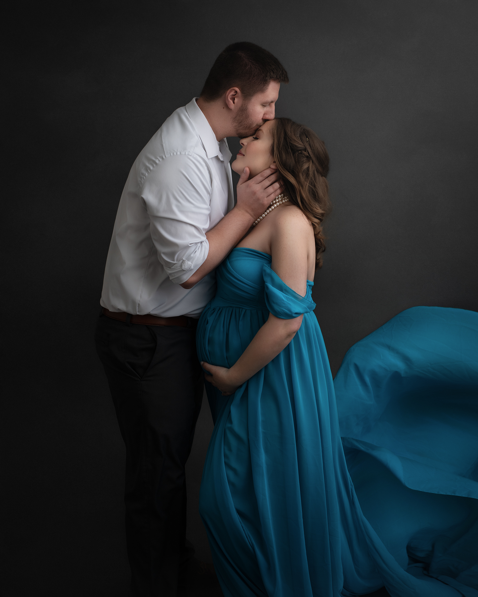 A pregnant woman stands in a studio wearing a long blue maternity gown while her husband kisses her forehead st louis baby shower venues